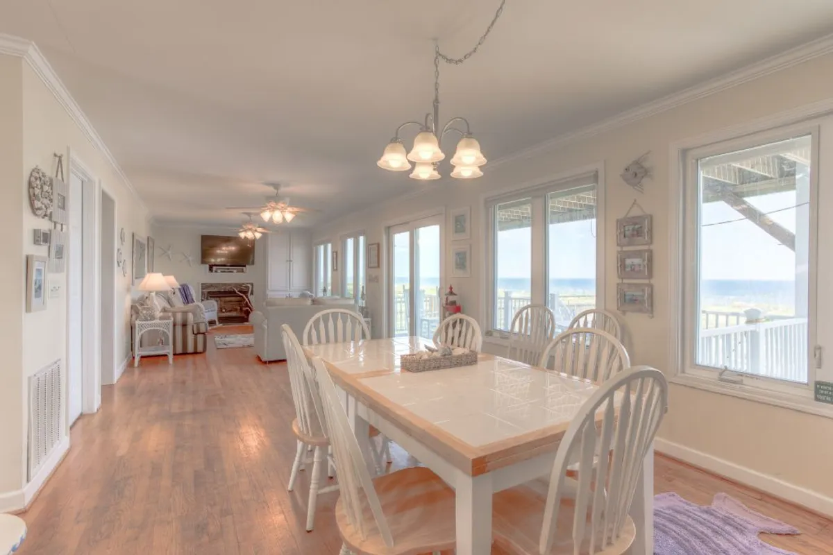 A warm dining area with a large wooden table surrounded by comfortable chairs, positioned before wide windows that offer glimpses of the ocean. The setting is ideal for family dinners, celebrations, and conversations, with soft, welcoming light enhancing the communal feel.