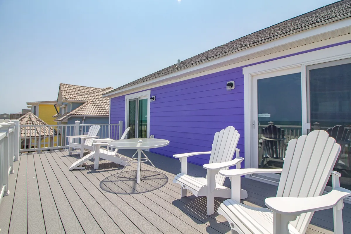 This exterior scene showcases a laid-back porch outfitted with white Adirondack chairs around a circular glass table, set against a colorful purple wall. The setting is casual and cheerful, ideal for relaxed conversation or enjoying the view, with hints of the beach environment beyond.