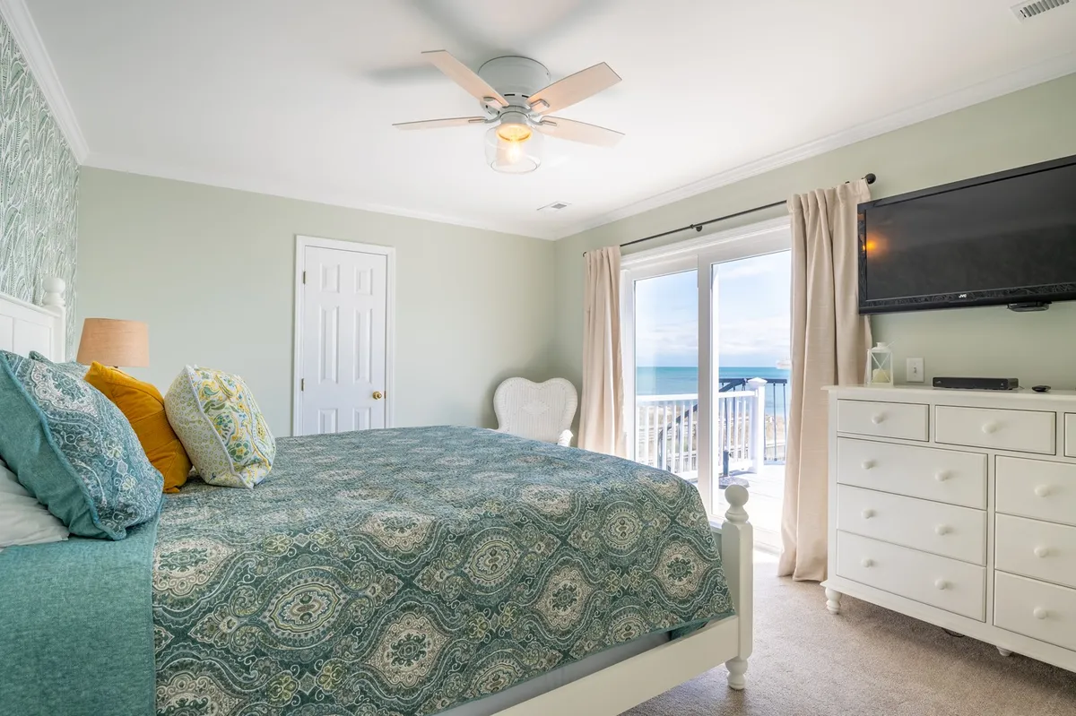 A bedroom scene featuring a bed, balcony doors, dresser, and television, with natural light streaming in, suggesting direct access to a private balcony and inviting guests to take in the view or enjoy morning coffee outside.