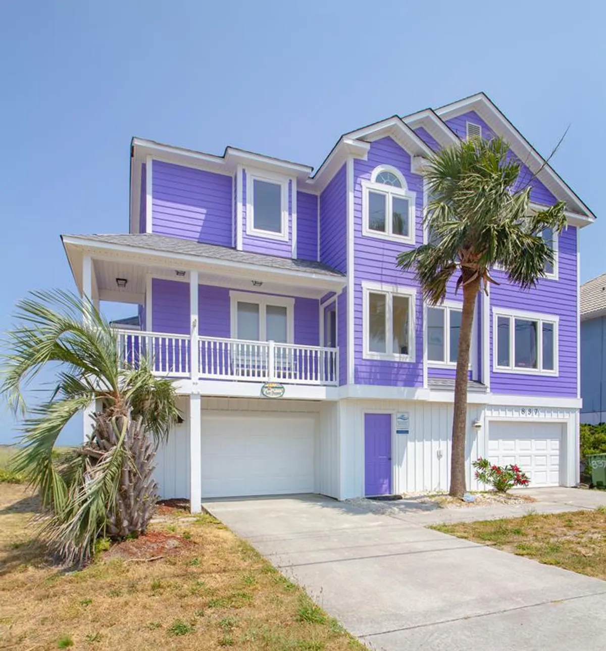 A cheerful exterior shot of the home's purple façade, featuring large windows, a balcony, palm trees, and a welcoming front yard. The balcony, visible and accessible, hints at more opportunities for relaxation and socializing with a view.