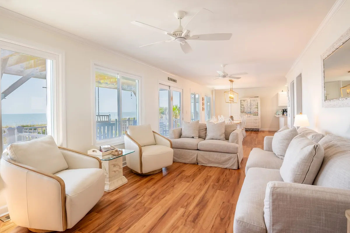 A spacious living room featuring large windows that frame an expansive view of the ocean. The room offers plush, comfortable seating, a decorative mirror, and light wood flooring, all bathed in natural light. The inviting atmosphere encourages relaxation and shared moments with loved ones.
