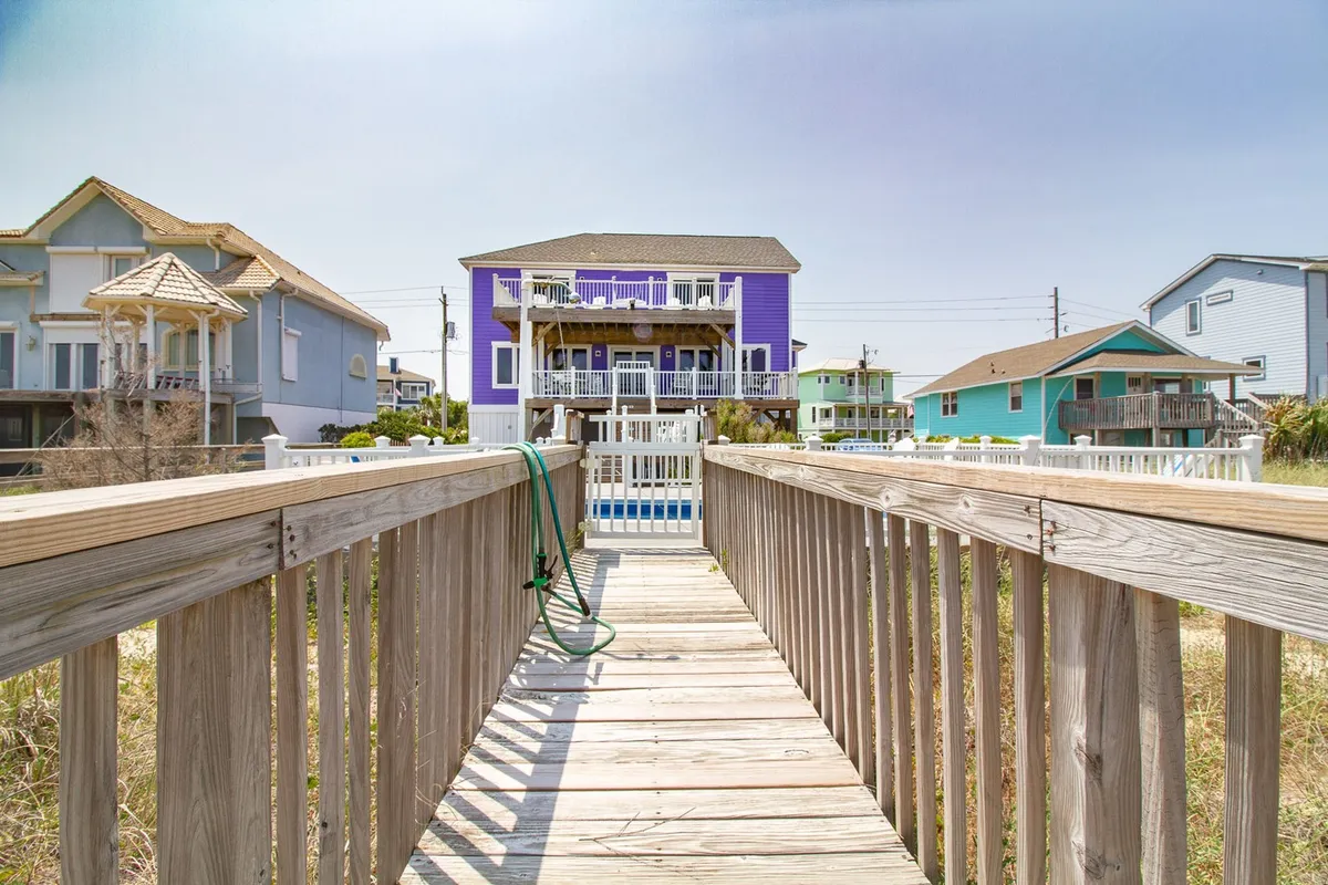 The image features a colorful exterior scene, including a clear blue pool, wooden walkway, and charming surrounding beach houses. The wooden boardwalk leads from the pool area toward the sand, creating a sense of easy movement between spaces.