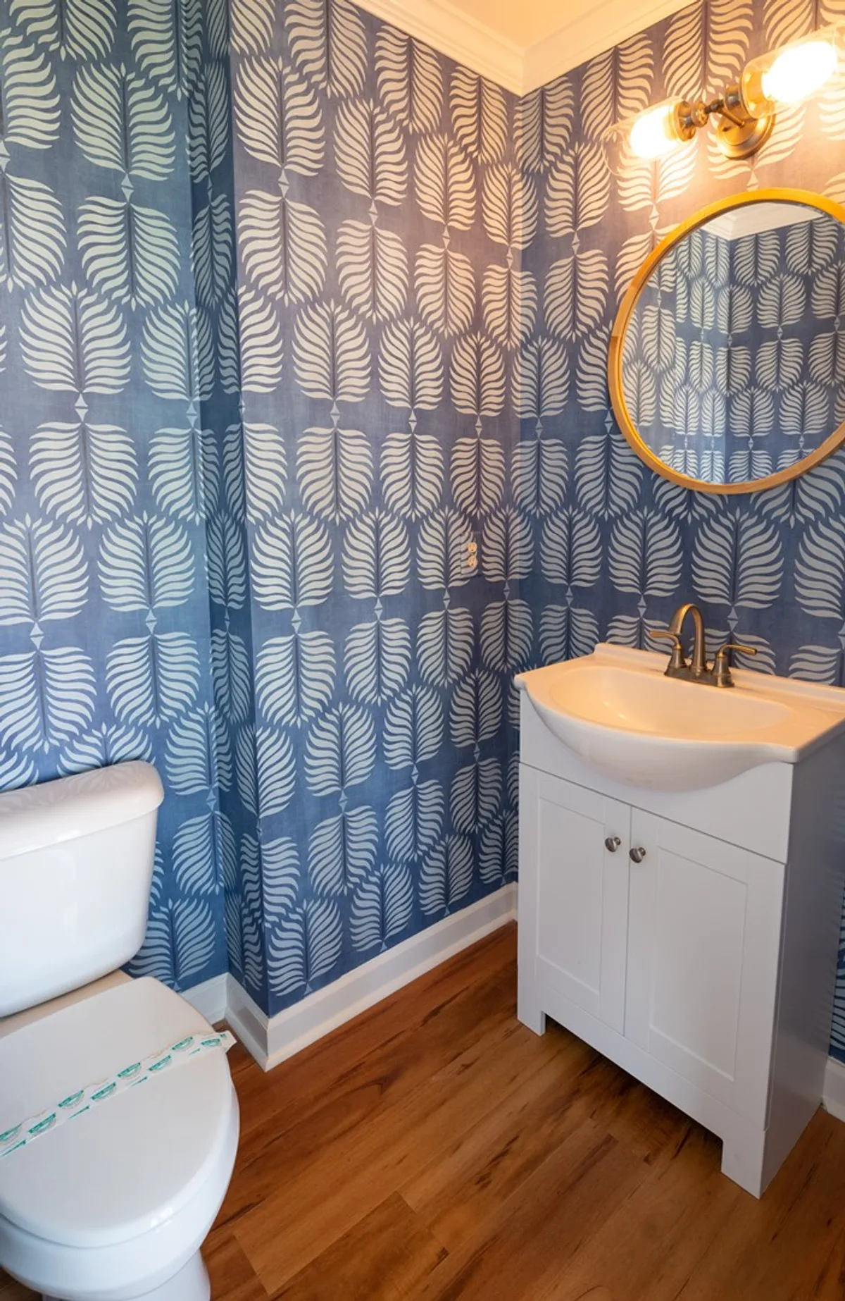 A stylish bathroom with uplifting blue patterned wallpaper, a round mirror above a modern sink and faucet, and warm wooden flooring. The space feels both refreshing and serene—a perfect spot to start or end the day.