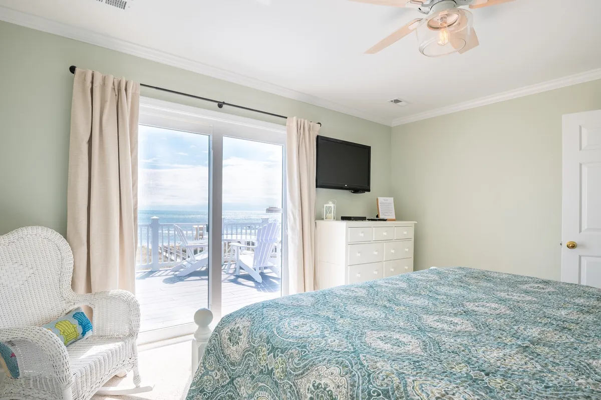 A serene bedroom with patterned bedding, a wicker chair, and a large sliding glass door leading directly to the deck, inviting guests to step outside and embrace the coastal environment.