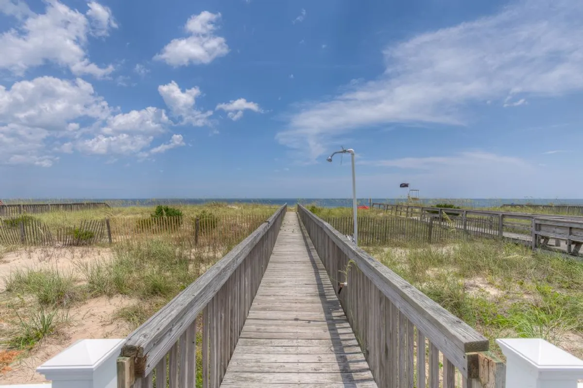 A wooden boardwalk leads over dune grass straight to the soft sand of Kure Beach, with the ocean and a cloudy sky as a stunning backdrop. The scene captures the sense of direct, private beachfront access, with plenty of space for beach games and sandcastle building.