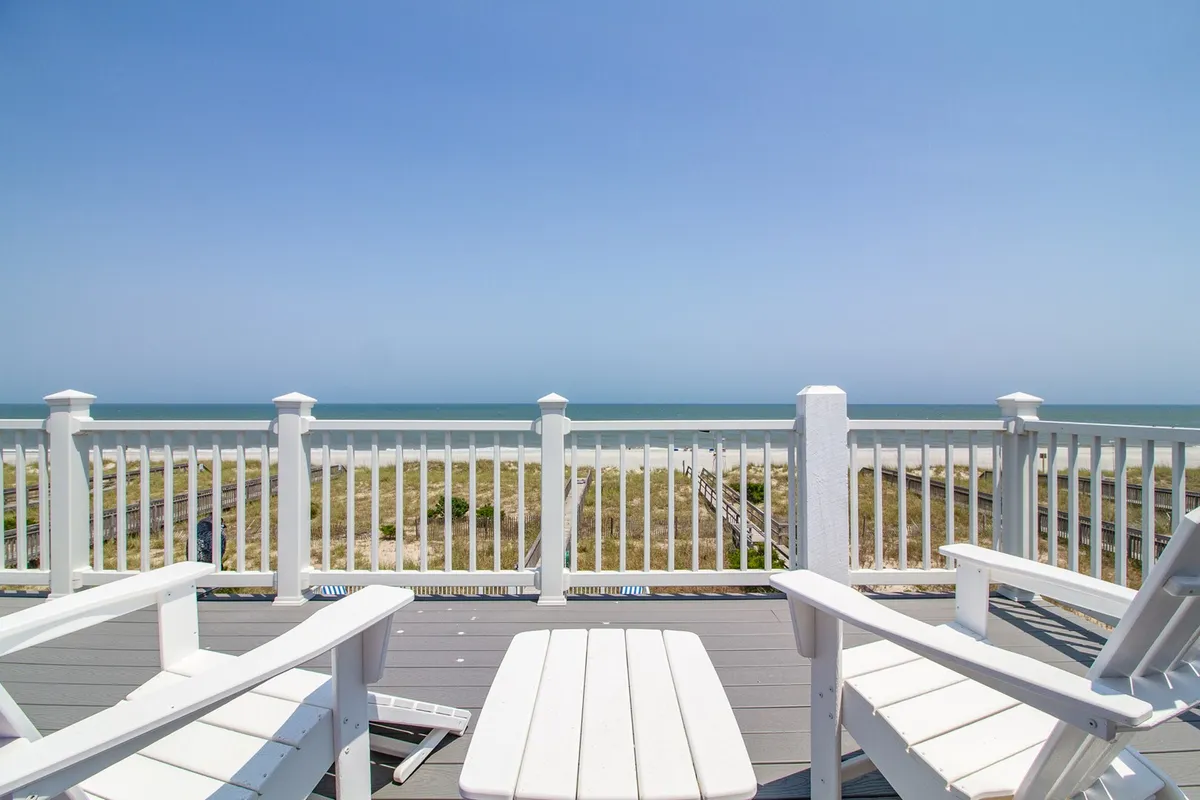A covered oceanfront porch with classic white Adirondack chairs and a sturdy railing, all overlooking the shimmering Atlantic. The porch invites guests to relax, enjoy the sea breeze, and soak in unobstructed ocean views—ideal for morning coffee or sunset gatherings.