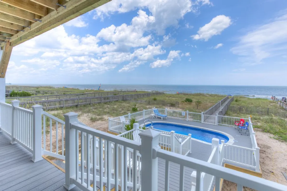 This image captures a wide view of the private pool, deck, and ocean beyond. The pool shimmers under open sky, bordered by the deck with ample seating and the seamless transition to beach sands visible in the frame. The setup makes it easy to imagine going from a swim in the pool to a walk on the sand.