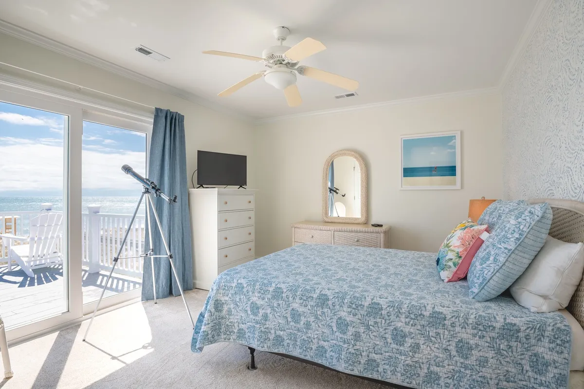 A spacious bedroom with a blue patterned cover on the bed, large windows that frame a stunning ocean view, a telescope positioned for stargazing, and a dresser with a mirror. The room is filled with natural light, offering a tranquil retreat.