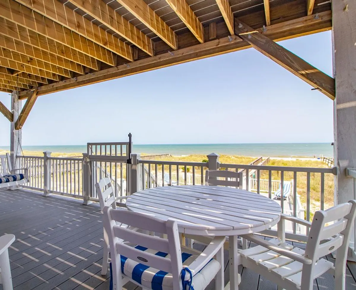 This exterior image captures a serene deck setting with a dining table and chairs, framed by a deck railing, and offering expansive views of the beach and ocean. The soft natural light and tranquil vibe suggest an ideal spot for shared meals, morning coffee, or simply enjoying the panoramic coastline.