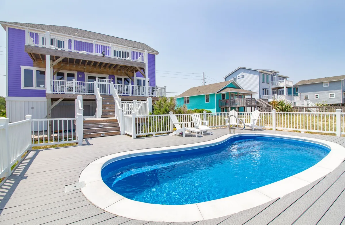 A vibrant purple oceanfront house with sprawling white decks overlooks a sparkling private pool surrounded by lush greenery. The scene is set for relaxing swims, sun-soaked afternoons, and joyful gatherings—all framed by the breathtaking backdrop of Kure Beach.