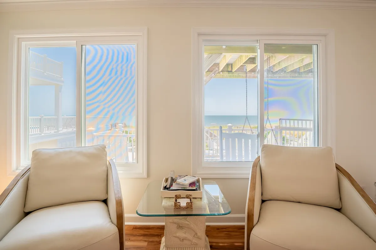 This image features a tranquil living space with large windows offering a prominent ocean view. Comfortable armchairs are arranged around a glass coffee table, inviting guests to relax, read, and enjoy the scenic setting.
