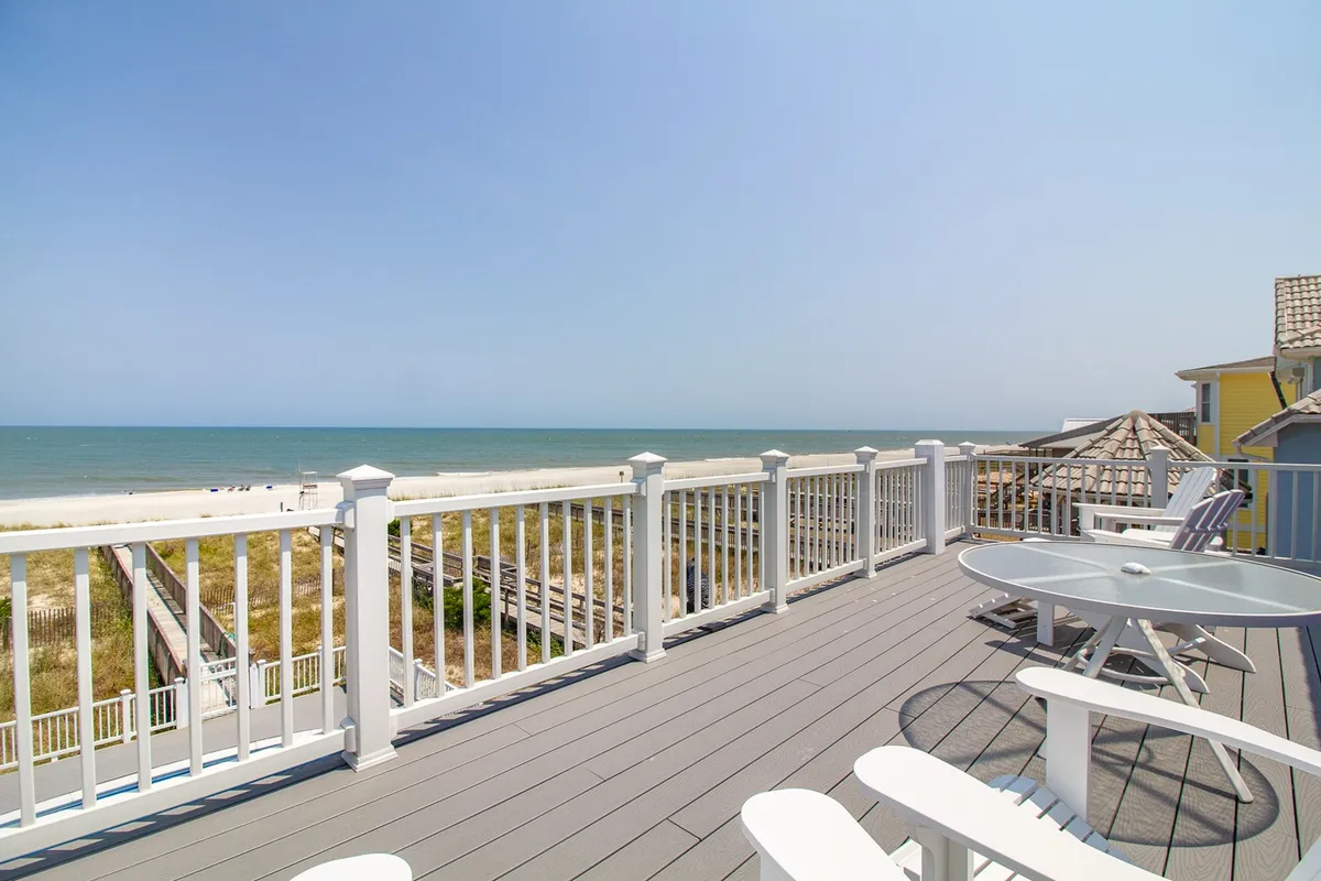 A spacious deck furnished for lounging and dining, overlooking the ocean with easy beach access visible beyond the railing. The tranquil vibe invites gatherings for meals, games, or simply soaking in the view as the sun sets.
