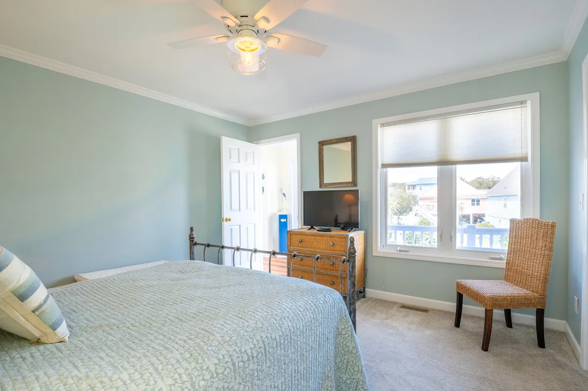 A peaceful bedroom with a bed covered in a light blue coverlet, a wooden dresser, a woven chair beside a bright window, and soft natural lighting. The atmosphere is calm and cozy, ideal for relaxation and private downtime.